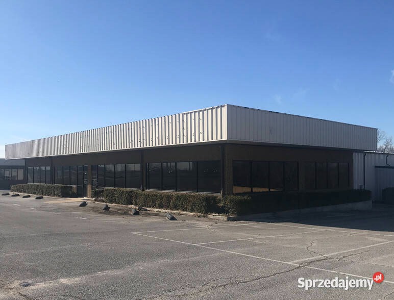 Niski budynek handlowy z płaskim dachem i rzędem okien, otoczony parkingiem i niskim żywopłotem.