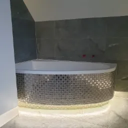 Narożna wanna obudowana mozaiką w odcieniach szarości, podświetlona od dołu, z szarymi płytkami na ścianach w łazience na poddaszu.