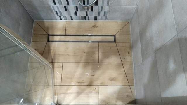 Wykończona kabina prysznicowa z odpływem liniowym, wyłożona płytkami imitującymi drewno w odcieniach beżu i brązu, z szarą mozaiką na ścianie i częściowo widoczną szklaną ścianką.