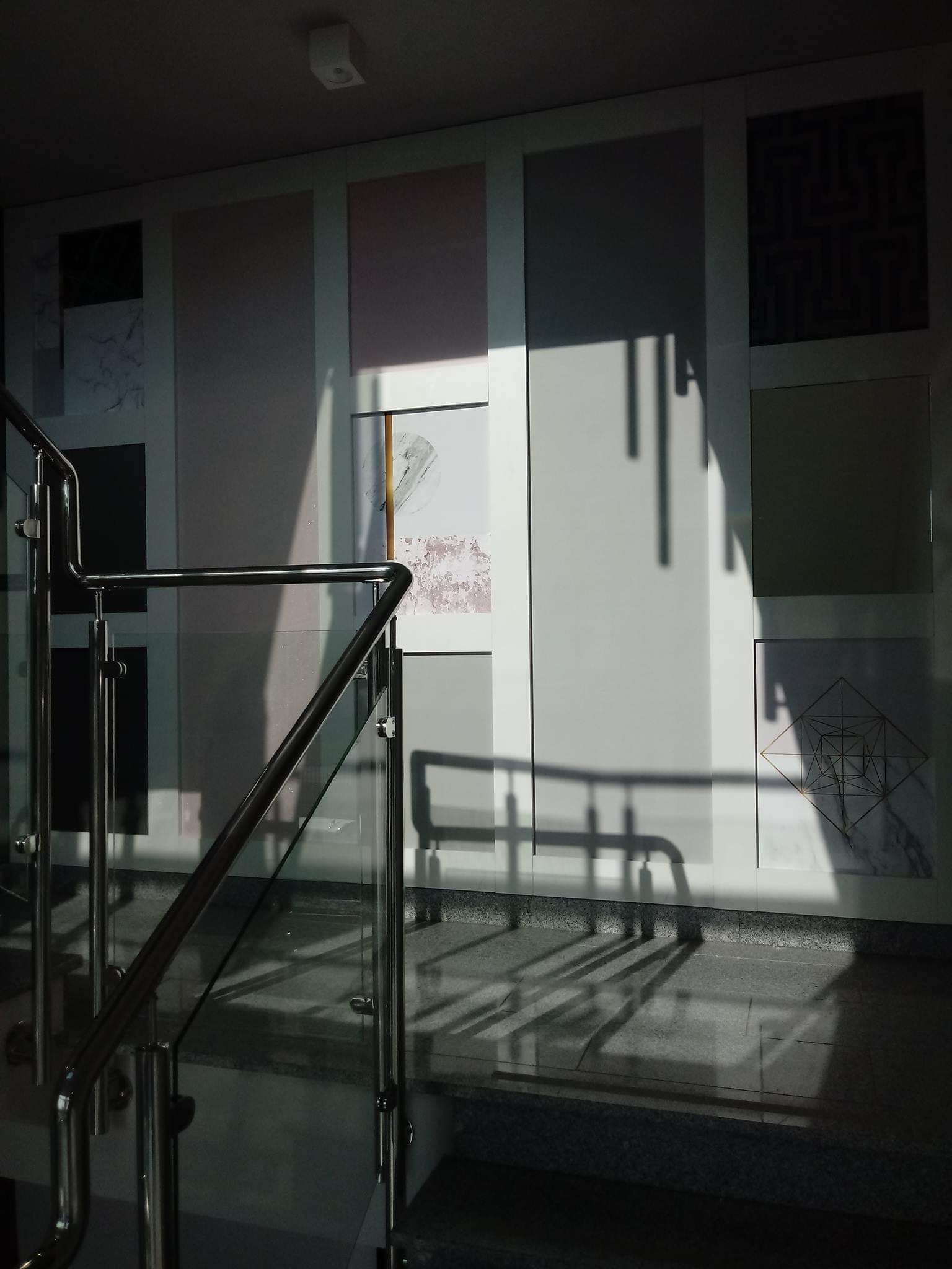 Klatka schodowa z szarą, granitową posadzką i nowoczesną balustradą ze stali nierdzewnej i szkła, ściana wykończona panelami z różnymi wzorami i fakturami, oświetlona padającym światłem słonecznym.
