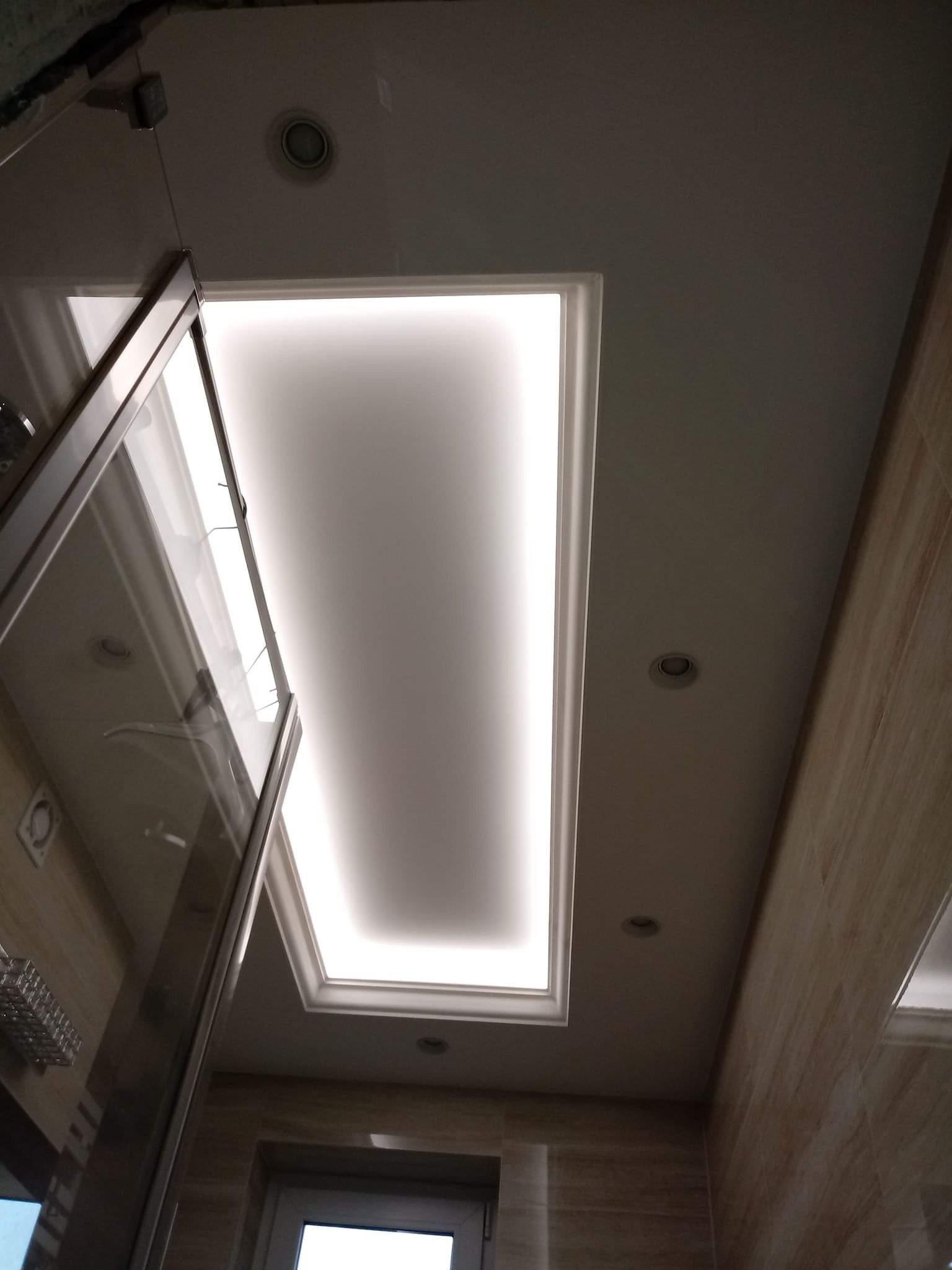 Podwieszany sufit w łazience z oświetleniem LED w kształcie prostokąta, obok kabina prysznicowa z chromowanymi profilami, widoczne oświetlenie punktowe i płytki imitujące drewno na ścianach.