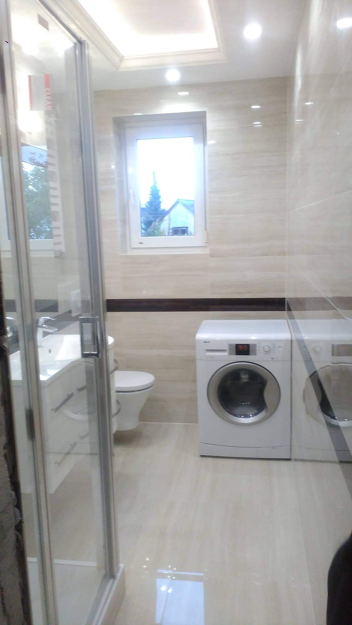 Jasna łazienka po remoncie z kabiną prysznicową, umywalką z szafką, toaletą i dwiema pralkami, wykończona płytkami imitującymi kamień z ciemnym pasem dekoracyjnym.