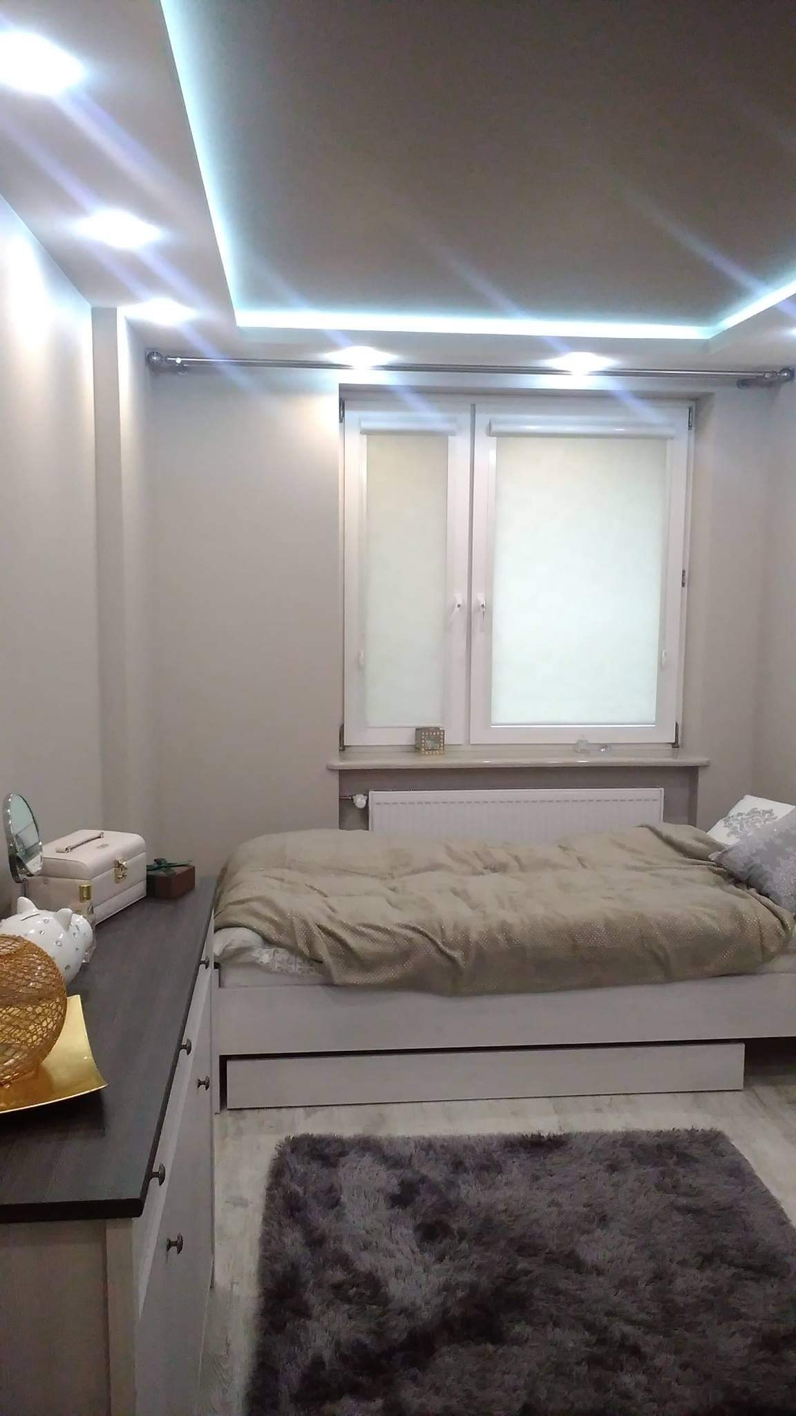 Jasny pokój z łóżkiem pod oknem, szary dywan, oświetlenie LED sufitowe, komoda z dekoracjami i lustrem.