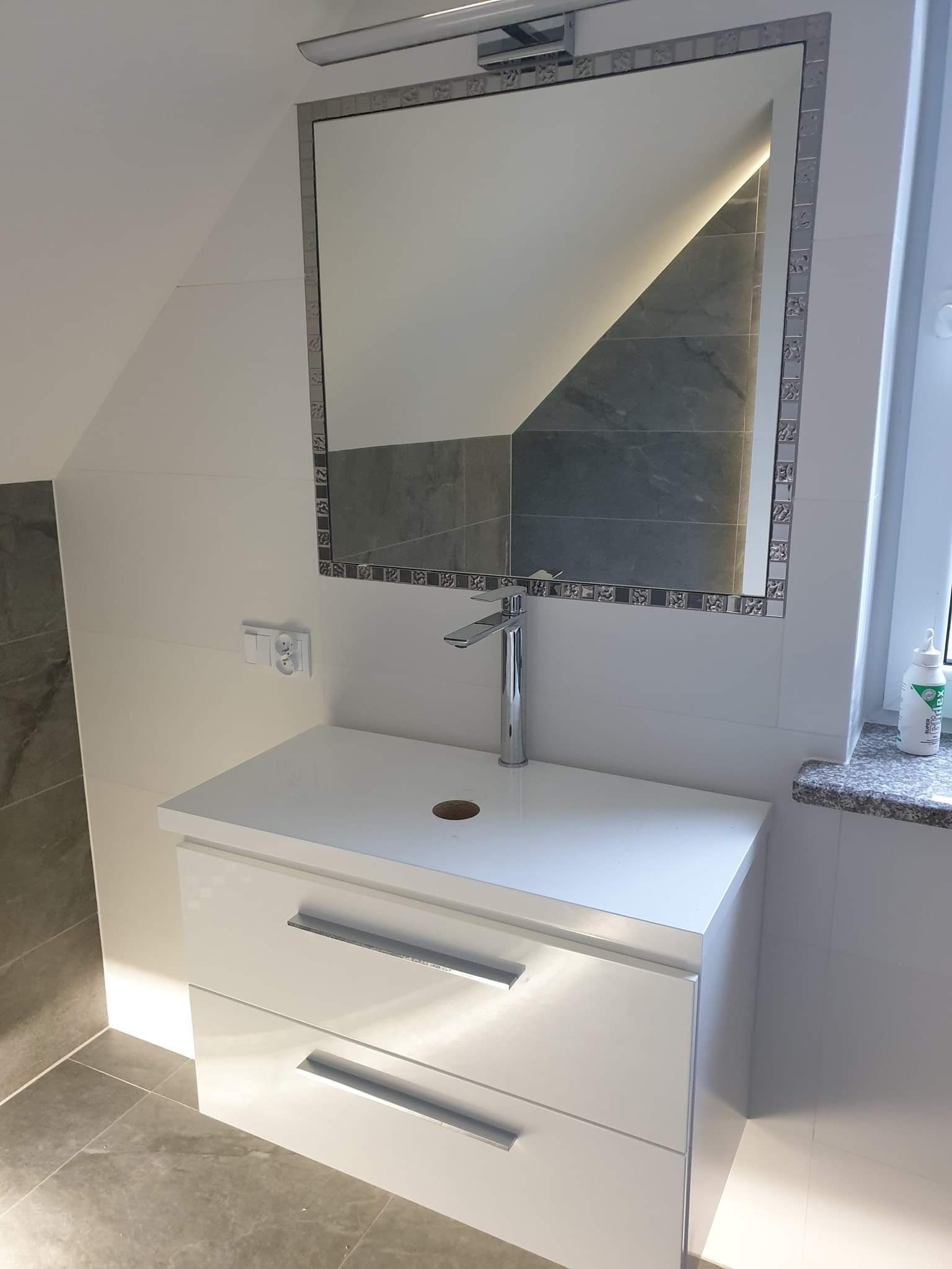 Biała szafka łazienkowa z umywalką i lustrem z ozdobną ramą w wykończonej łazience na poddaszu.