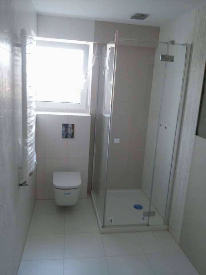 Nowoczesna łazienka z białą armaturą, kabiną prysznicową z transparentnego szkła i wiszącą toaletą, wykończona białymi płytkami z delikatnym wzorem.