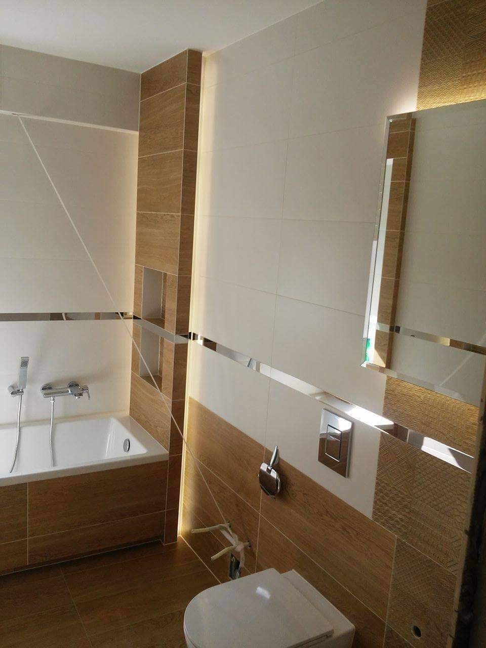 Nowoczesna łazienka z wanną obudowaną drewnopodobnymi panelami, białą ceramiką, lustrem z podświetleniem LED i akcentami w postaci chromowanych listew.