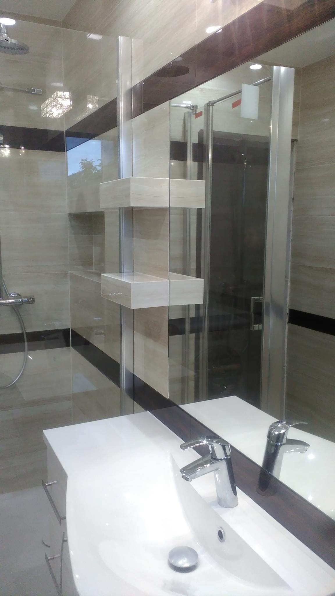 Nowoczesna łazienka z beżowymi płytkami, szklaną kabiną prysznicową i umywalką z chromowaną baterią, widoczne półki z jasnego drewna i dekoracyjny pas na ścianie.