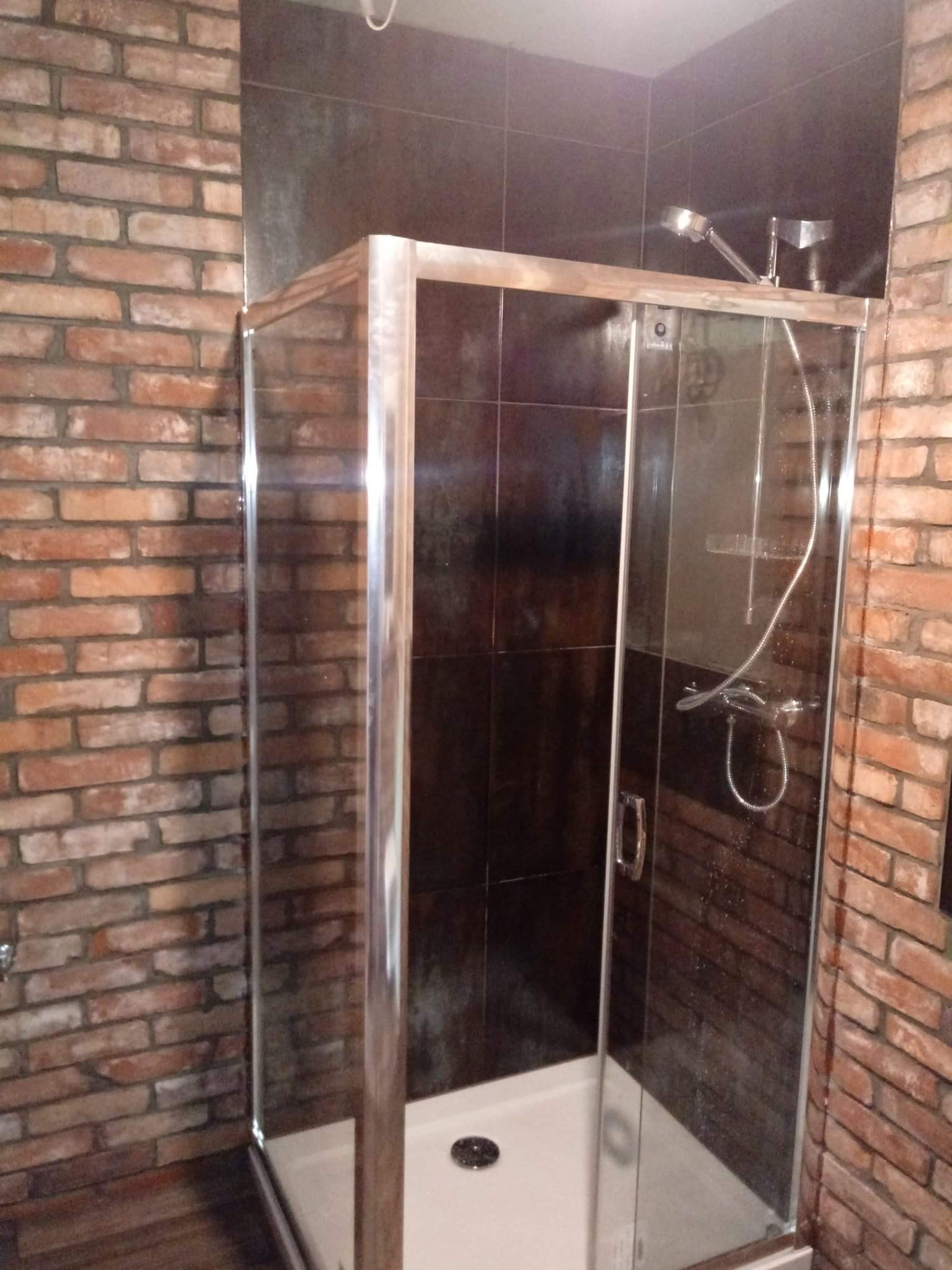 Kabina prysznicowa z przesuwnymi drzwiami, obudowana cegłą i ciemnymi płytkami, z widoczną słuchawką prysznicową i odpływem.