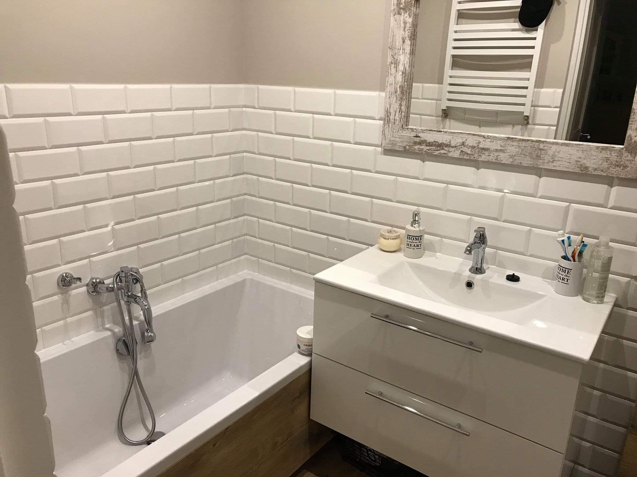 Biała łazienka z wanną i umywalką w szafce, wykończona białymi płytkami typu metro, z lustrem w ramie o efekcie postarzanego drewna.