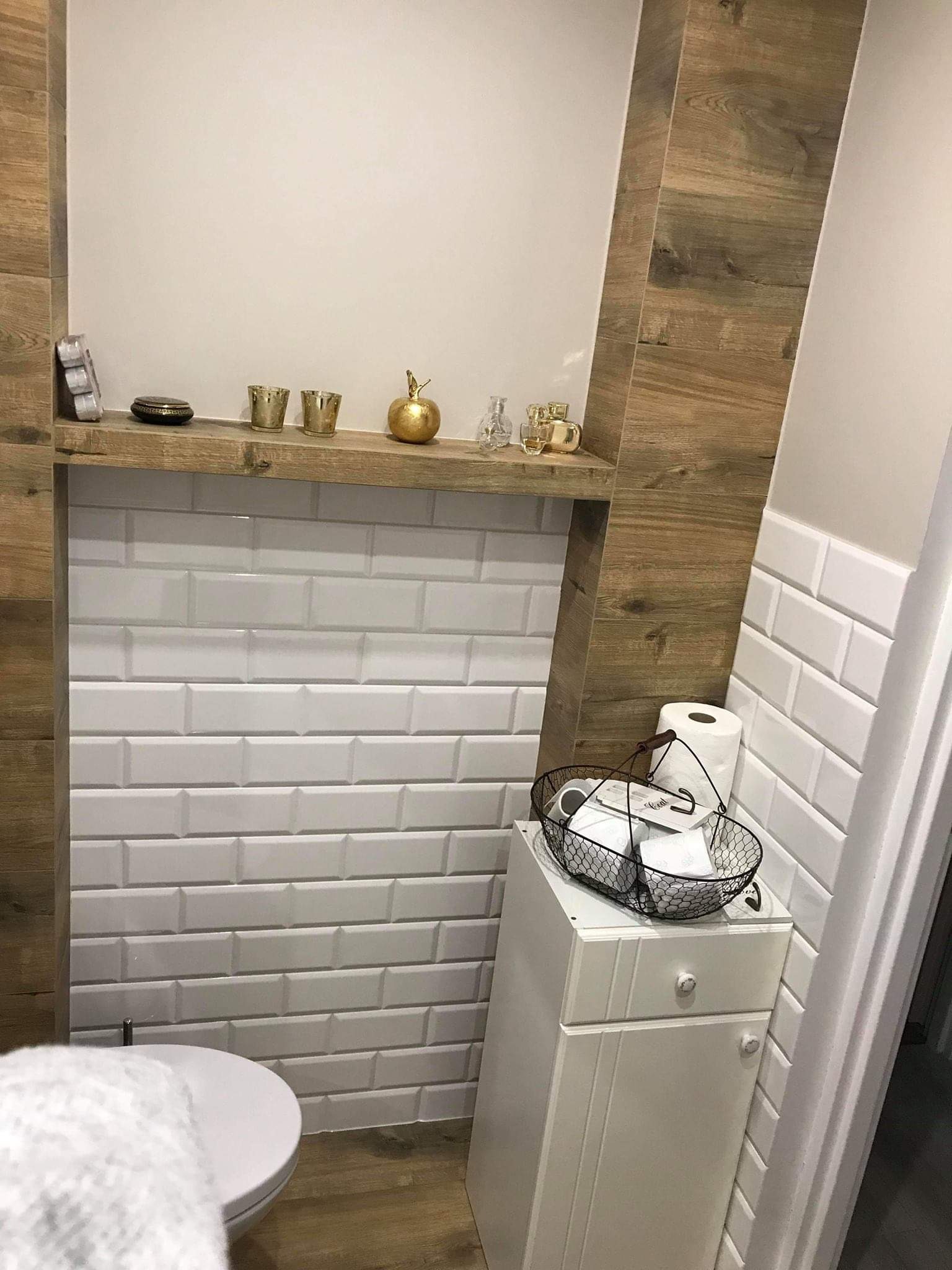 Nowoczesna toaleta z białymi, prostokątnymi płytkami przypominającymi cegiełki, drewnianym obramowaniem i półką z dekoracjami, uzupełniona białą szafką z koszykiem na akcesoria.
