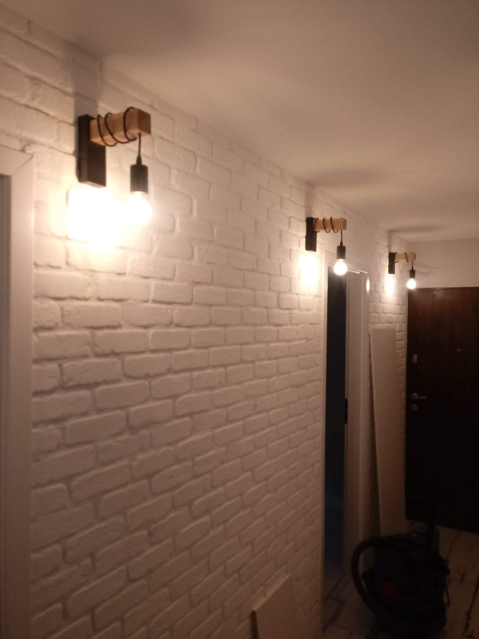 Korytarz z białymi ceglanymi ścianami i trzema industrialnymi lampami ściennymi z drewnianymi elementami.