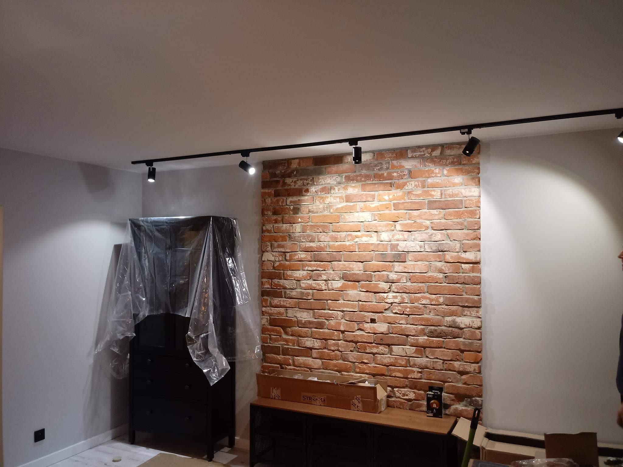 Wnętrze pokoju z odsłoniętą ceglaną ścianą, oświetleniem szynowym na suficie, zasłoniętą folią komodą i częściowo pomalowanymi ścianami.