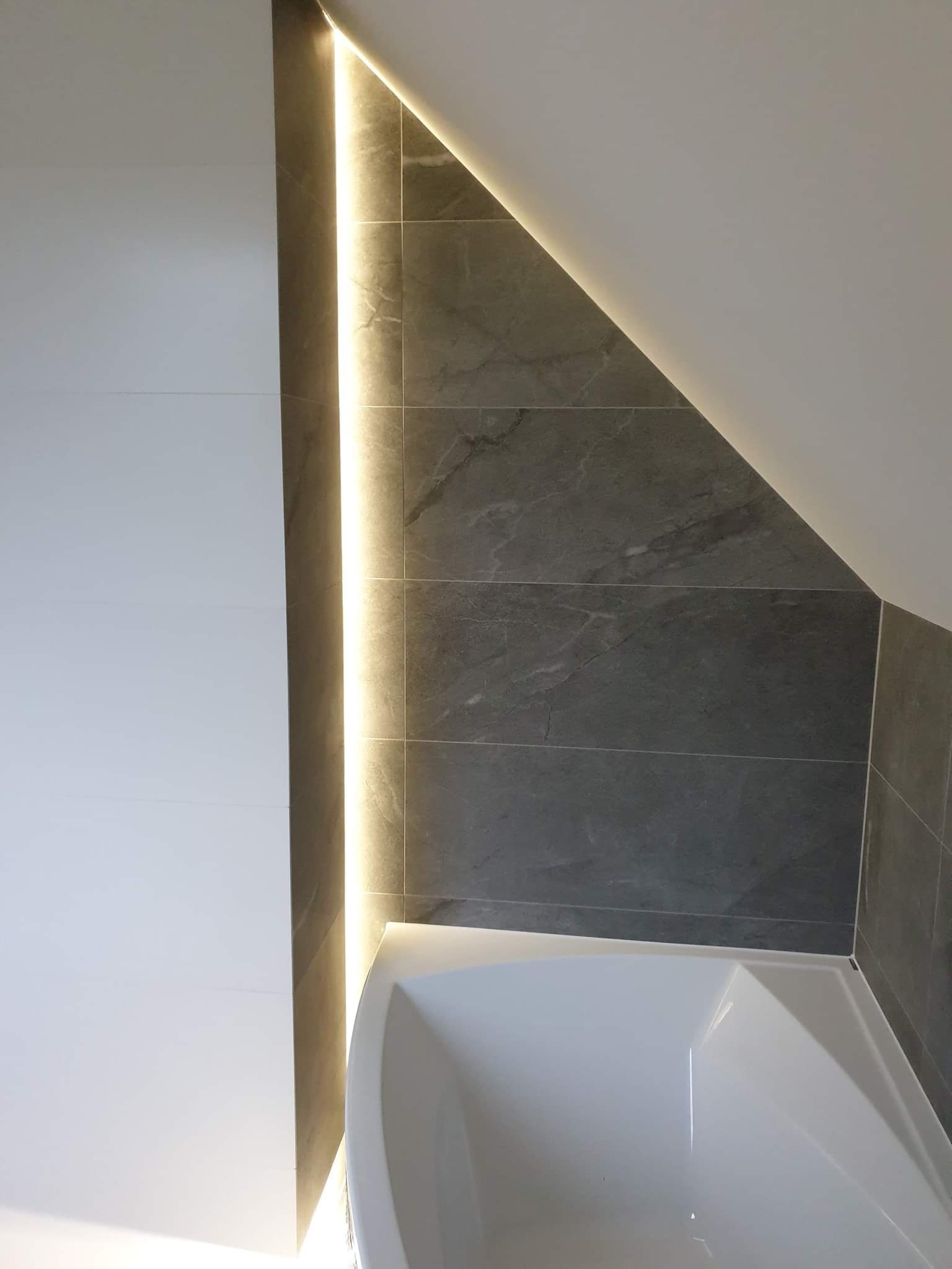 Nowoczesna łazienka z szarymi płytkami imitującymi kamień, podświetleniem LED w rogu i fragmentem białej wanny.