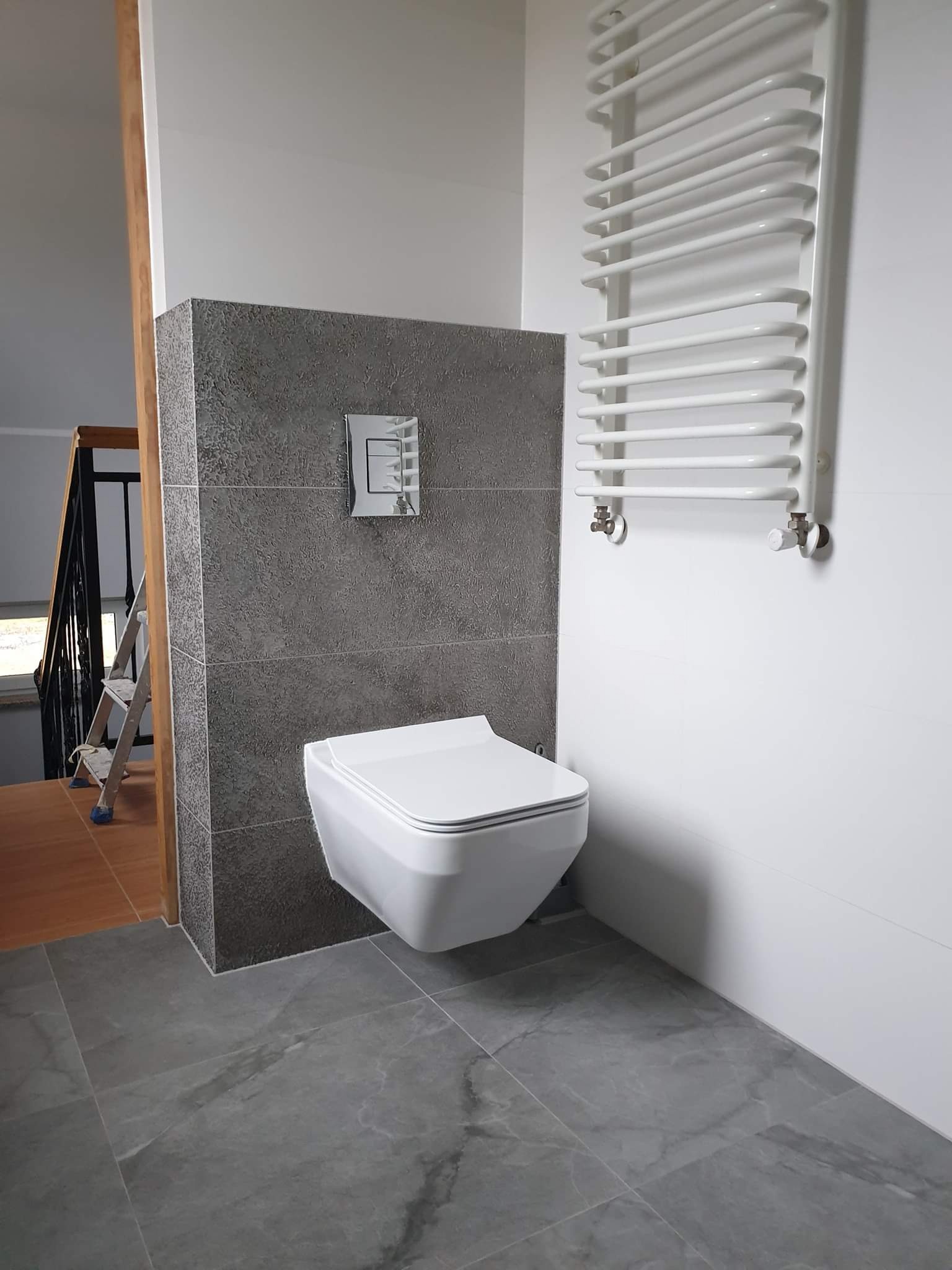 Nowoczesna toaleta podwieszana z szarą płytką imitującą kamień na ścianie, biały grzejnik drabinkowy i szare płytki podłogowe, widoczna drabina w tle.