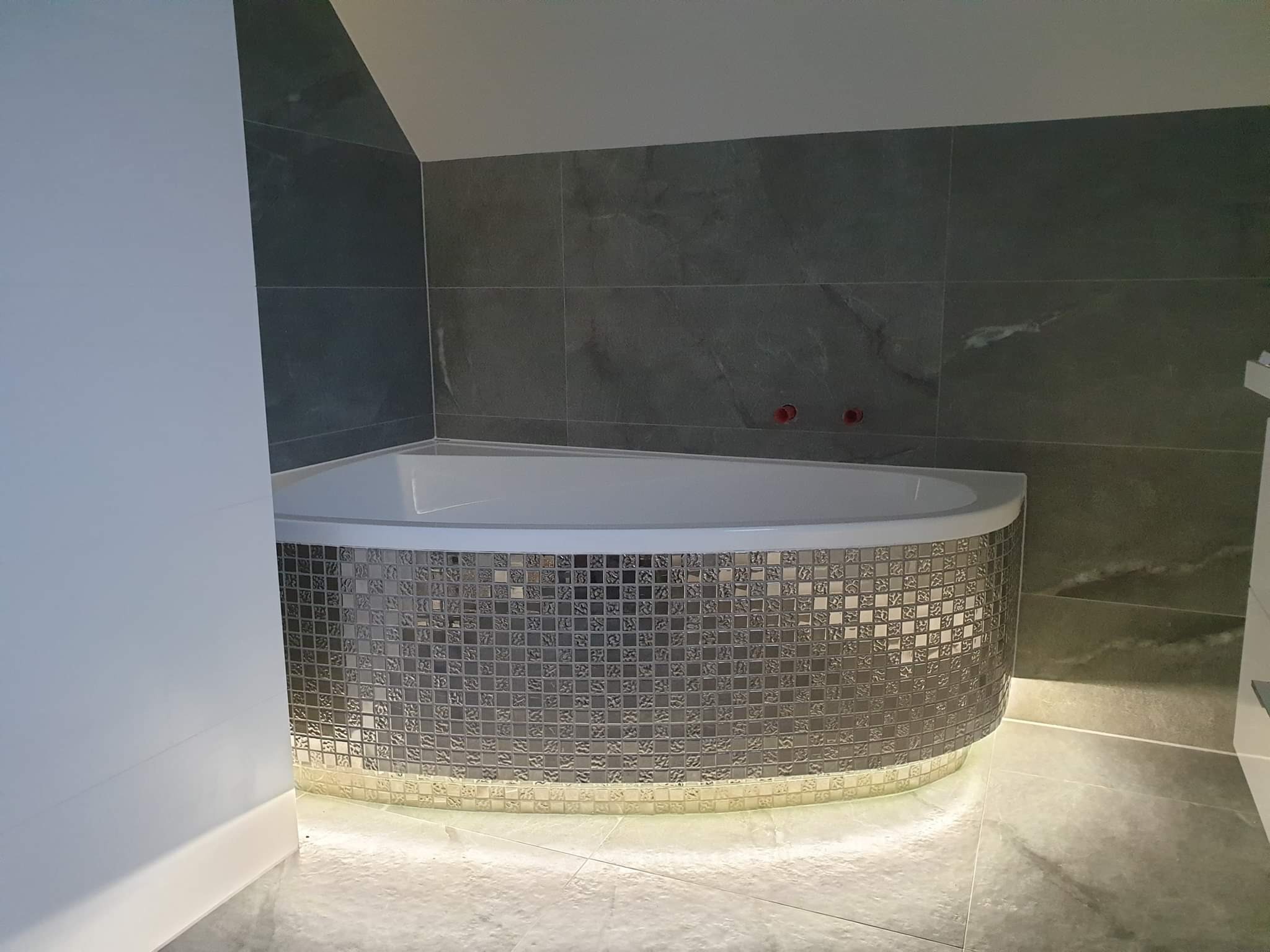 Narożna wanna obudowana mozaiką w odcieniach szarości, podświetlona od dołu, z szarymi płytkami na ścianach w łazience na poddaszu.