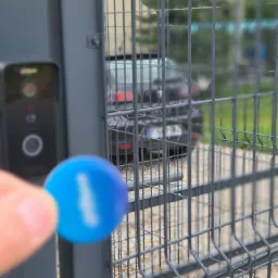 Zbliżenie na czytnik zbliżeniowy przy bramie wjazdowej z widoczną kartą dostępu i kamerą monitoringu, w tle zaparkowany samochód.