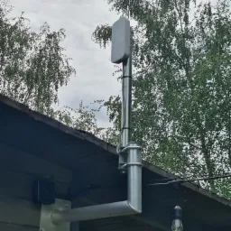 Zewnętrzna antena na metalowym maszcie przymocowana do budynku, z widoczną puszką przyłączeniową, na tle drzew i pochmurnego nieba.