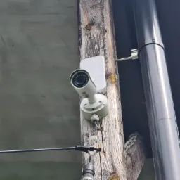 Biała kamera monitoringu zamontowana na szorstkim, drewnianym słupie z widocznymi sękami, obok rynny spustowej, z dekoracyjną girlandą żarówkową w tle.