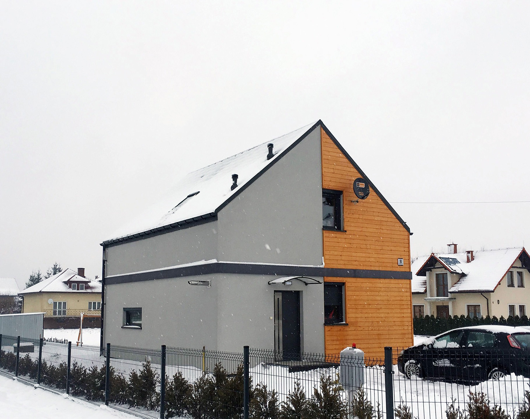 Nowoczesny dom szkieletowy w zimowej scenerii, z szarą elewacją po jednej stronie i drewnianą okładziną po drugiej, częściowo zasypany śniegiem, widoczny zza ogrodzenia.