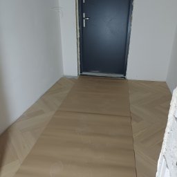 DAR BUD usługi ogólnobudowlane - Ułożona parkietowa podłoga w jodełkę, częściowo zakryta ochronną tekturą z odciskami butów, przy ciemnych drzwiach wejściowych i białych ścianach.