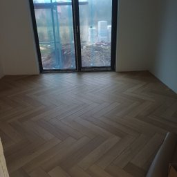 DAR BUD usługi ogólnobudowlane - Ułożona drewniana podłoga w jodełkę w jasnym pomieszczeniu z dużym oknem balkonowym. Widoczne materiały budowlane za oknem.