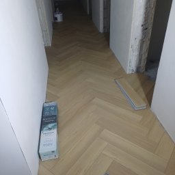 DAR BUD usługi ogólnobudowlane - Ułożona podłoga w jodełkę w korytarzu, widoczne niedokończone ściany i paczka paneli podłogowych Aqua Profi. Jasne panele tworzą wzór jodełki.