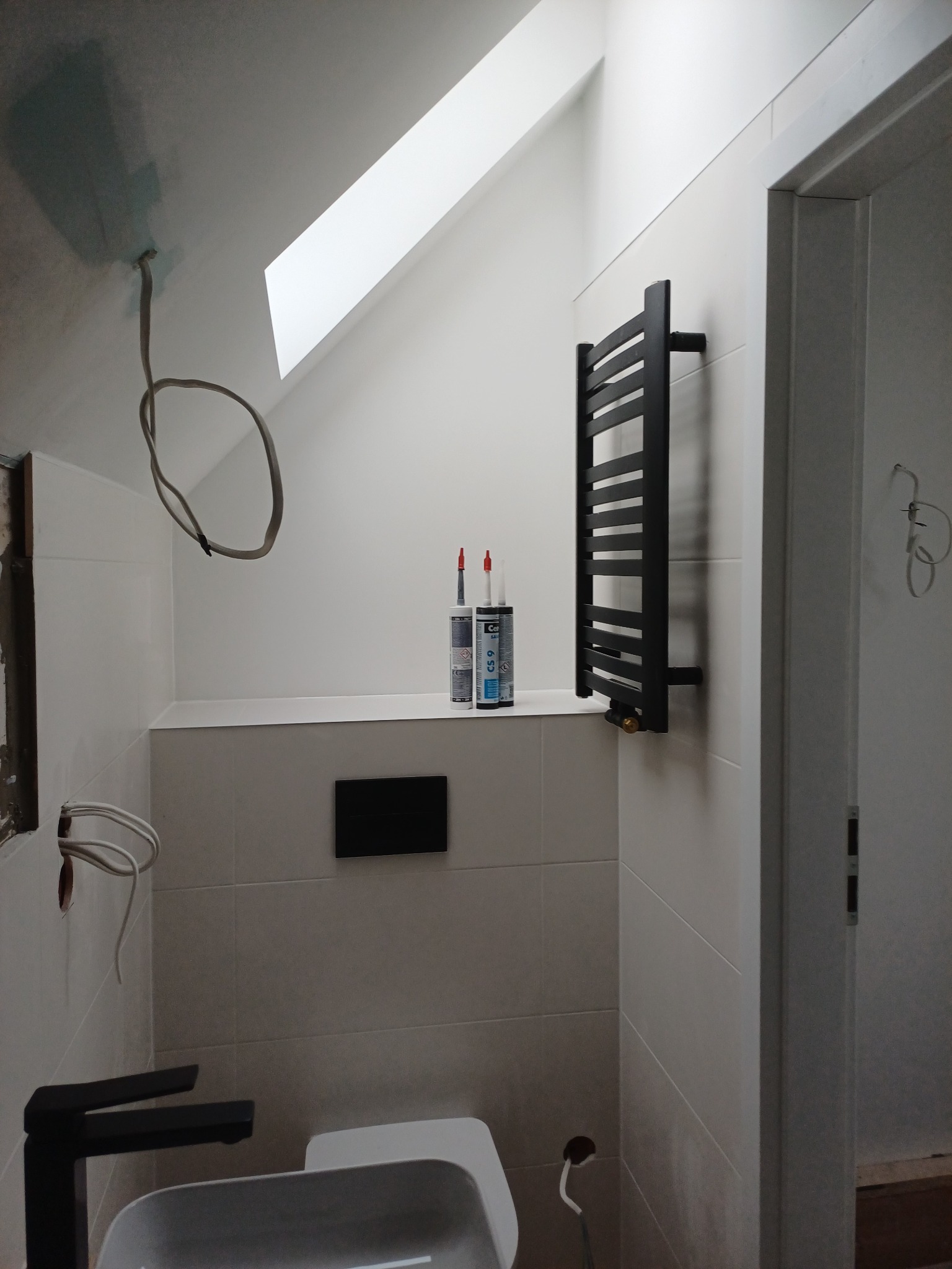 Łazienka w trakcie wykończenia: białe płytki, czarny grzejnik drabinkowy, częściowo zamontowana armatura, widoczne przewody i materiały budowlane. Skos dachu z oknem.