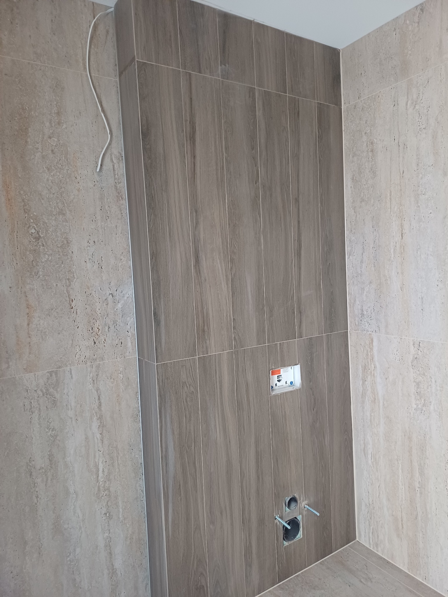 Wykończona ściana w łazience z brązowymi płytkami imitującymi drewno, widoczne otwory na armaturę i wystający kabel.