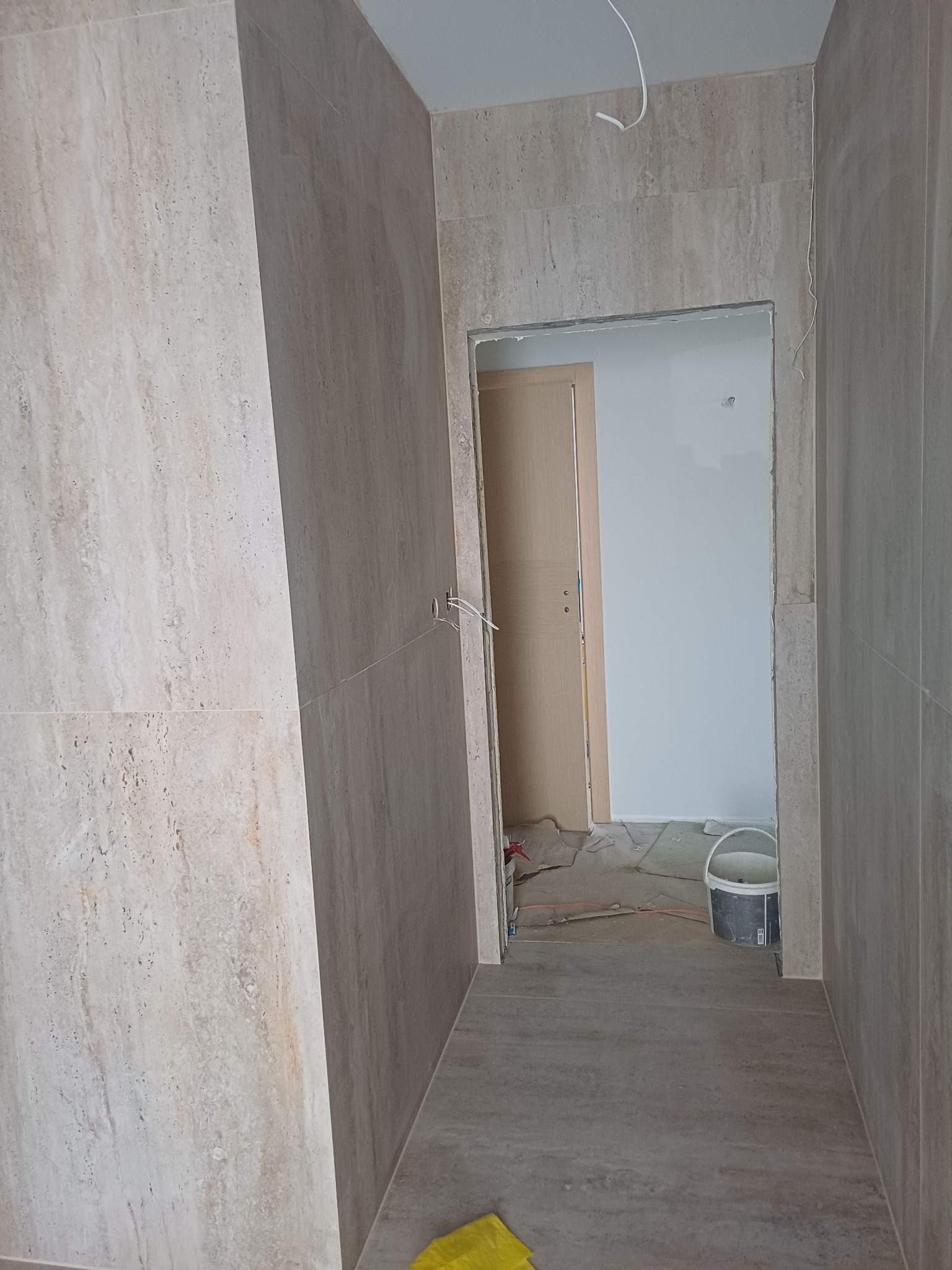 Wnętrze łazienki w trakcie wykańczania: beżowe płytki na ścianach i podłodze, widoczne narzędzia i wiadro, surowy stan wykończenia, przewody elektryczne wystające ze ściany.