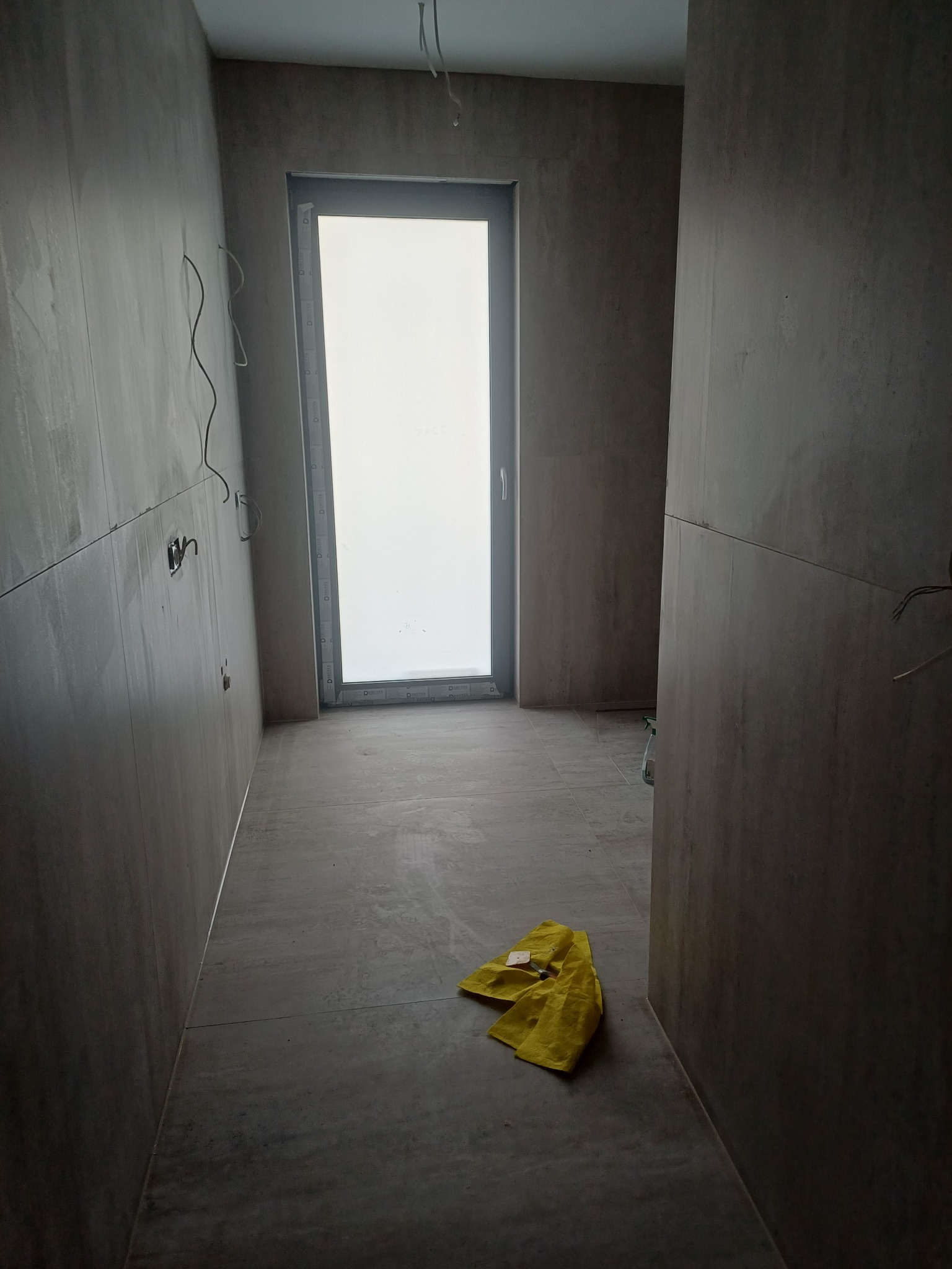 Wykończona łazienka z szarymi płytkami, widoczne gniazdka elektryczne i okno. Na podłodze leży żółta ścierka. Minimalistyczny design, surowe wykończenie.