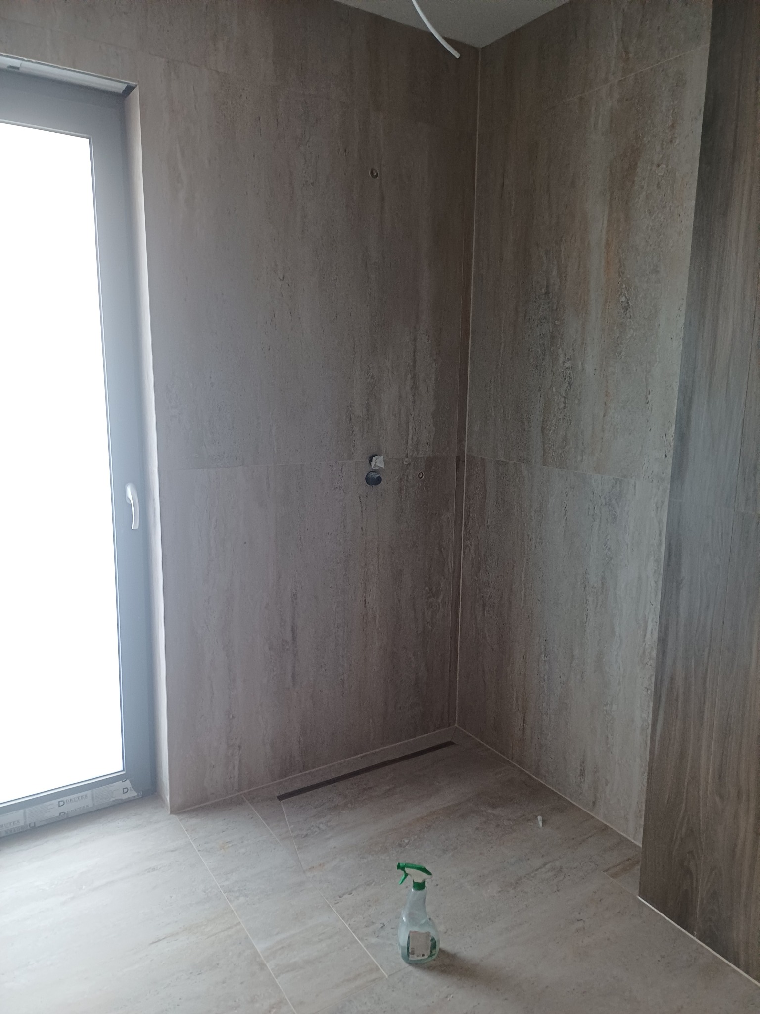 Wykończona kabina prysznicowa z odpływem liniowym i beżowymi płytkami imitującymi kamień. Widoczne przyłącza wodne i okno. Na podłodze butelka ze spryskiwaczem.