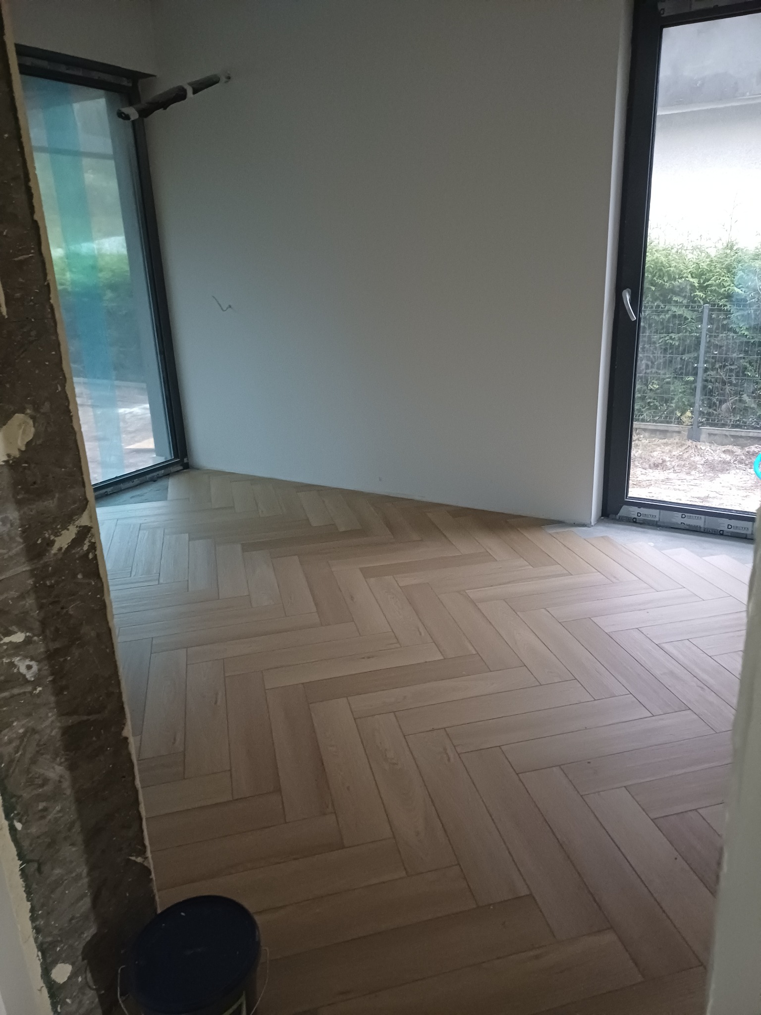 Jasna, drewniana podłoga ułożona w jodełkę w nowym wnętrzu z dużymi oknami. Widoczny fragment ściany w stanie surowym i puszka farby.