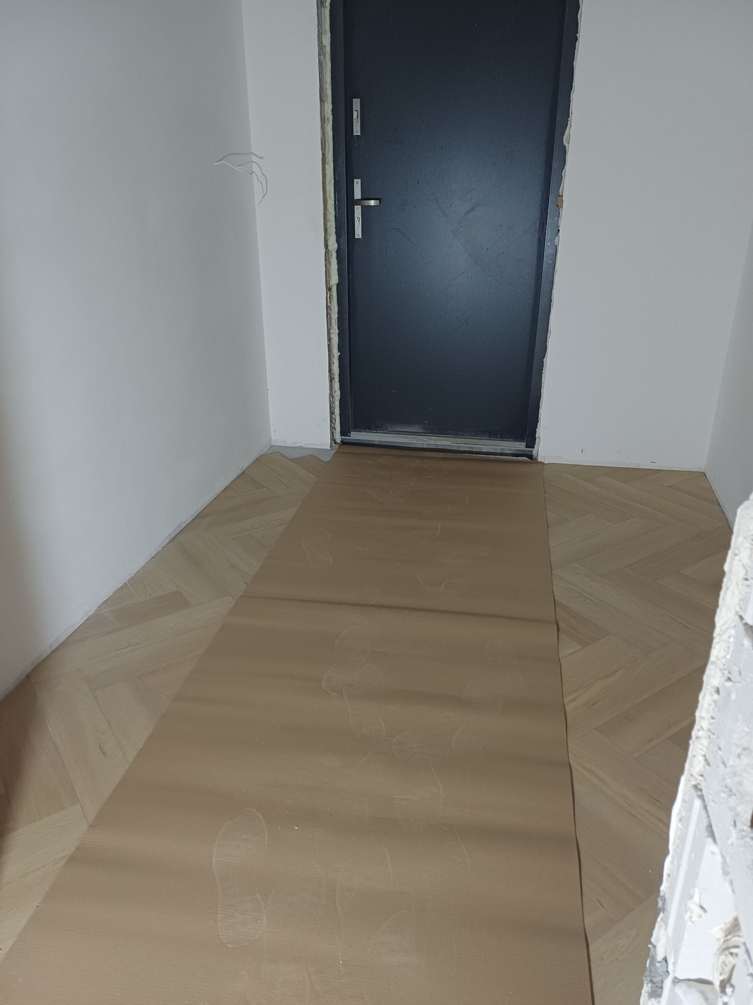 Ułożona parkietowa podłoga w jodełkę, częściowo zakryta ochronną tekturą z odciskami butów, przy ciemnych drzwiach wejściowych i białych ścianach.