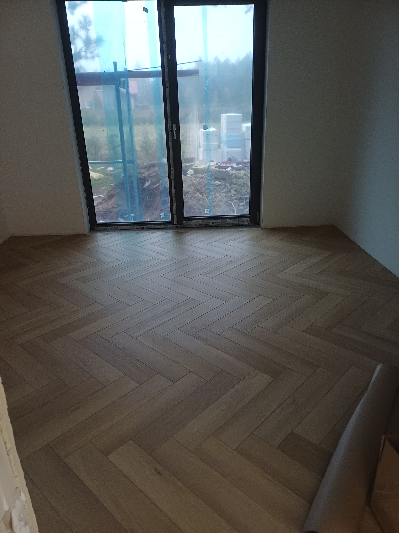 Ułożona drewniana podłoga w jodełkę w jasnym pomieszczeniu z dużym oknem balkonowym. Widoczne materiały budowlane za oknem.