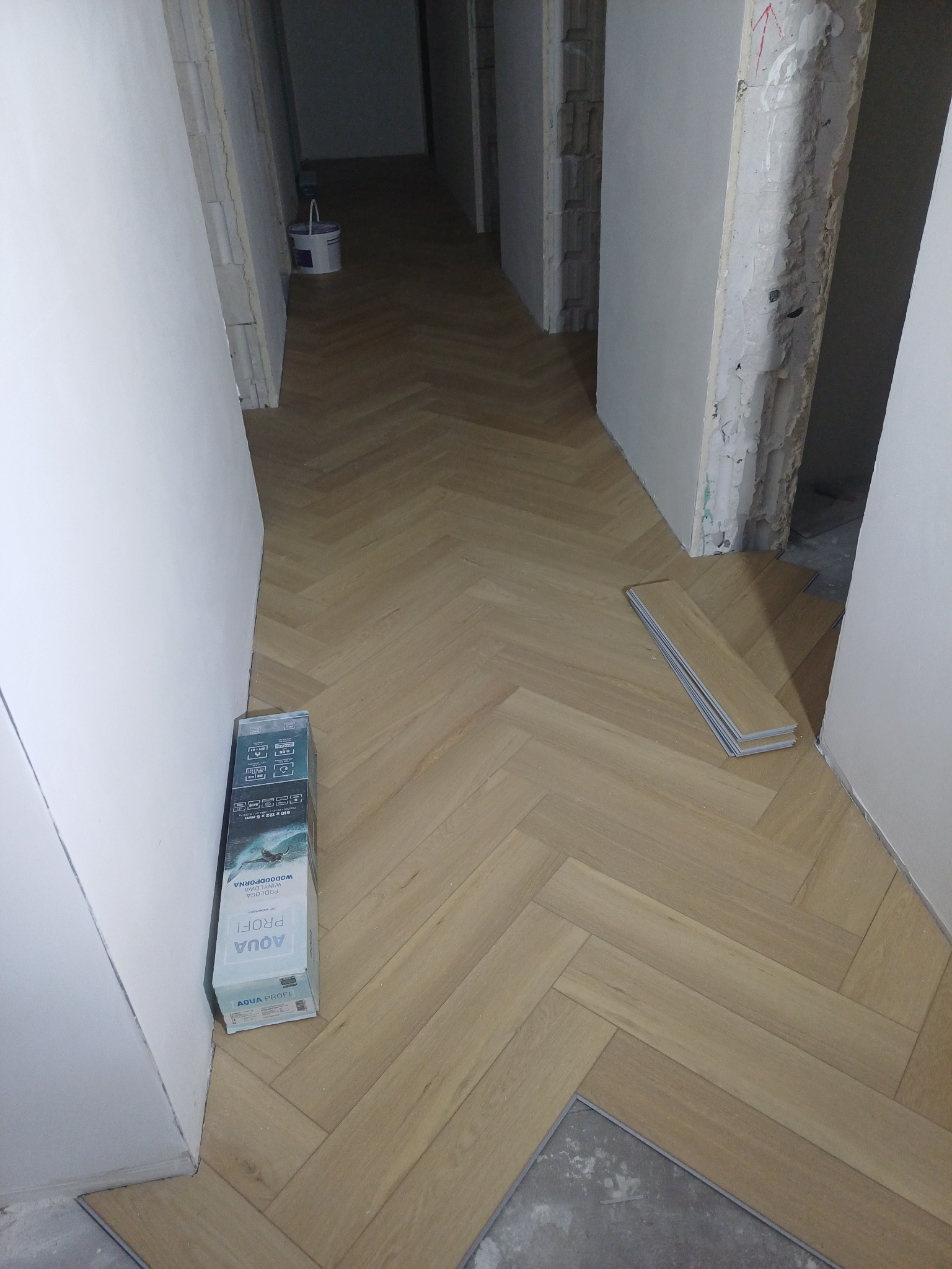 Ułożona podłoga w jodełkę w korytarzu, widoczne niedokończone ściany i paczka paneli podłogowych Aqua Profi. Jasne panele tworzą wzór jodełki.