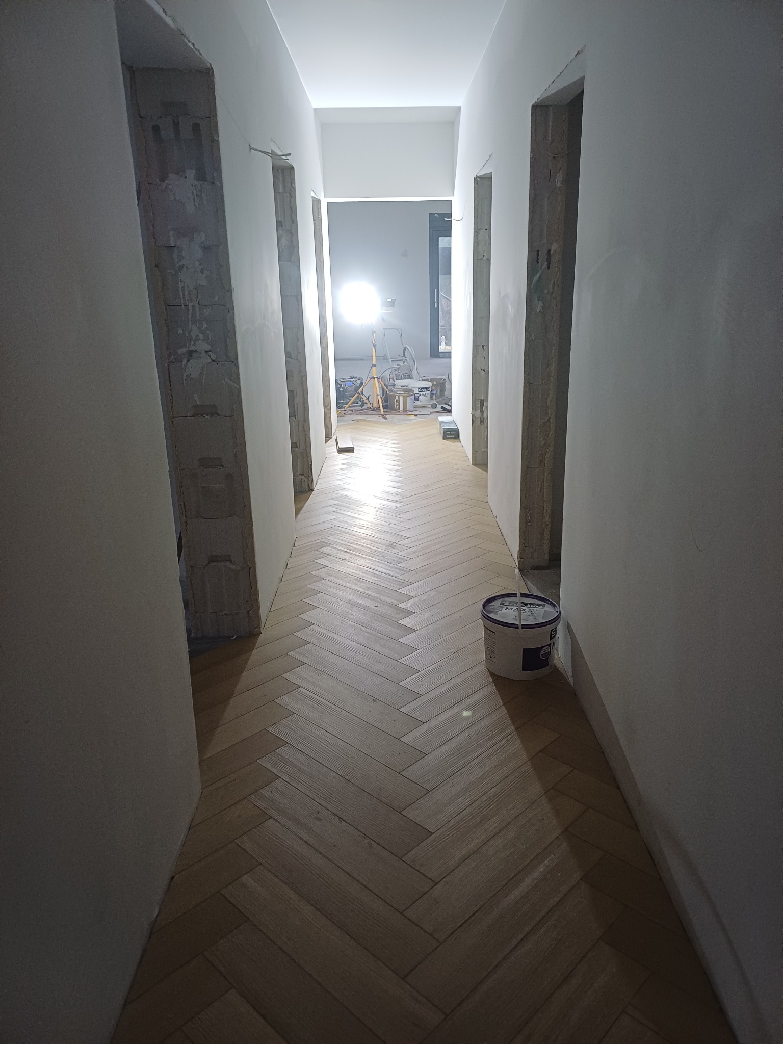 Korytarz w trakcie remontu: świeżo położona drewniana podłoga w jodełkę, ściany przygotowane do malowania, widoczne surowe wnęki drzwiowe i wiadro z farbą.