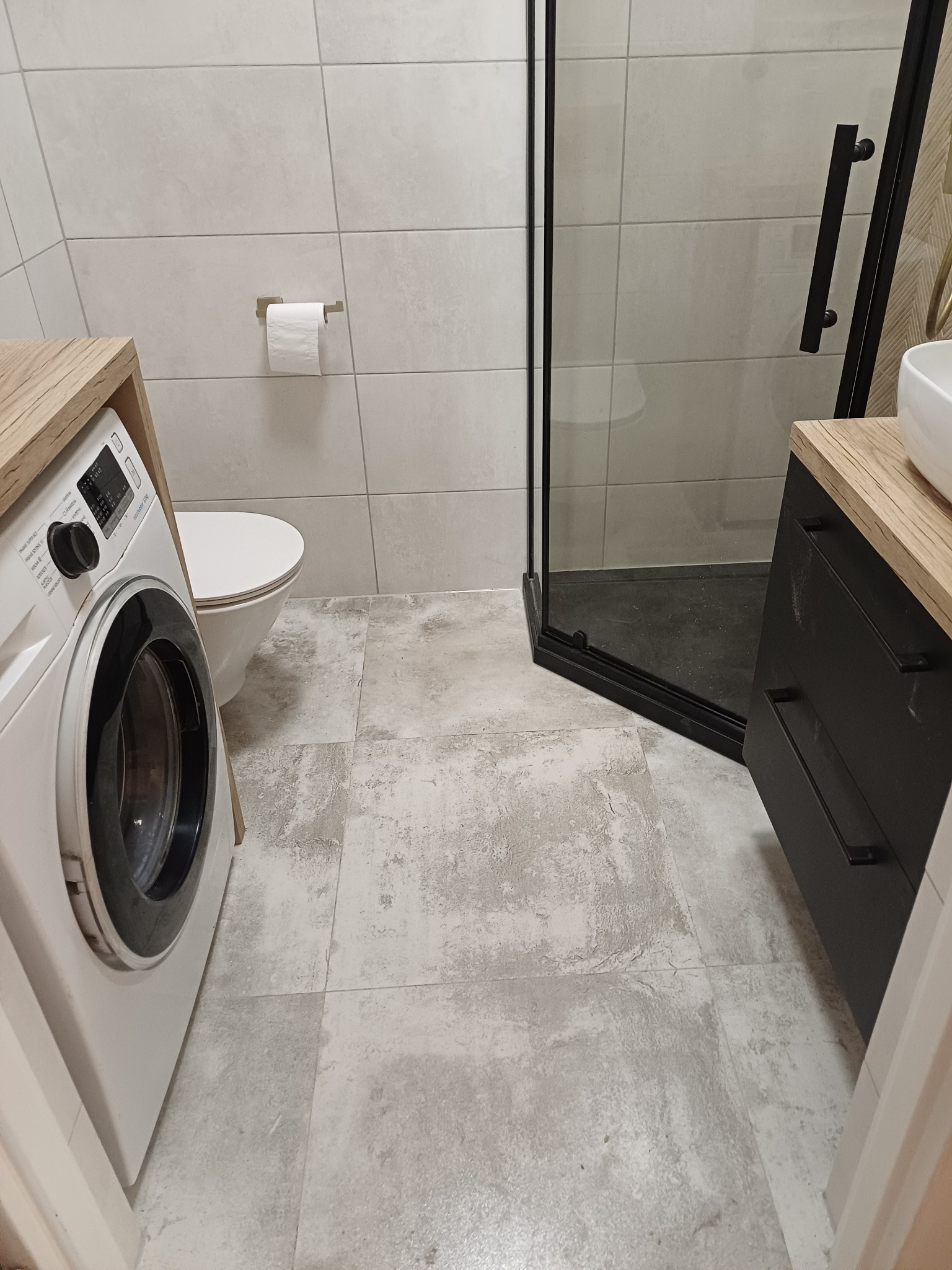 Nowoczesna łazienka z kabiną prysznicową typu walk-in, pralką i toaletą. Jasne płytki imitujące beton, czarne akcenty i drewniane blaty. Minimalistyczny design.