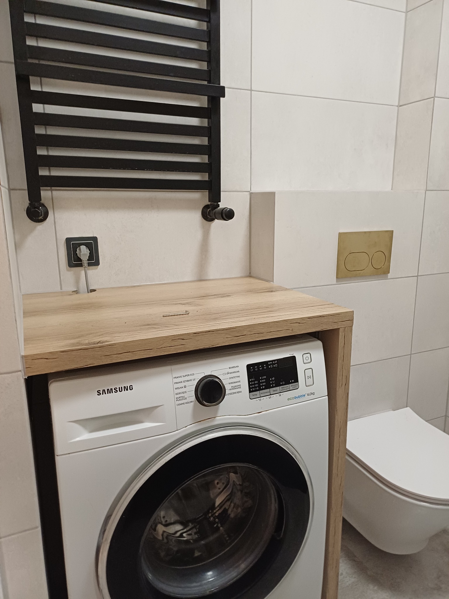 Nowoczesna łazienka z pralką Samsung zabudowaną pod drewnianym blatem, grzejnik drabinkowy i podwieszana toaleta. Minimalistyczny design, płytki w jasnych odcieniach.