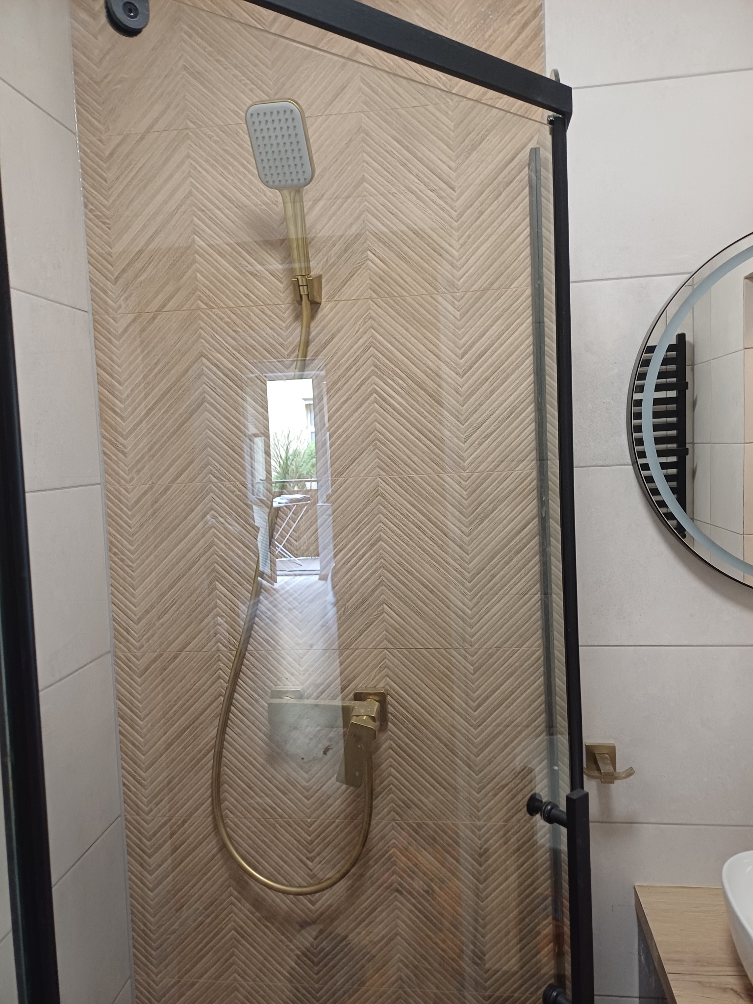 Kabina prysznicowa z deszczownicą w łazience wykończonej płytkami w jodełkę. Widoczne czarne ramy kabiny i okrągłe lustro z podświetleniem.