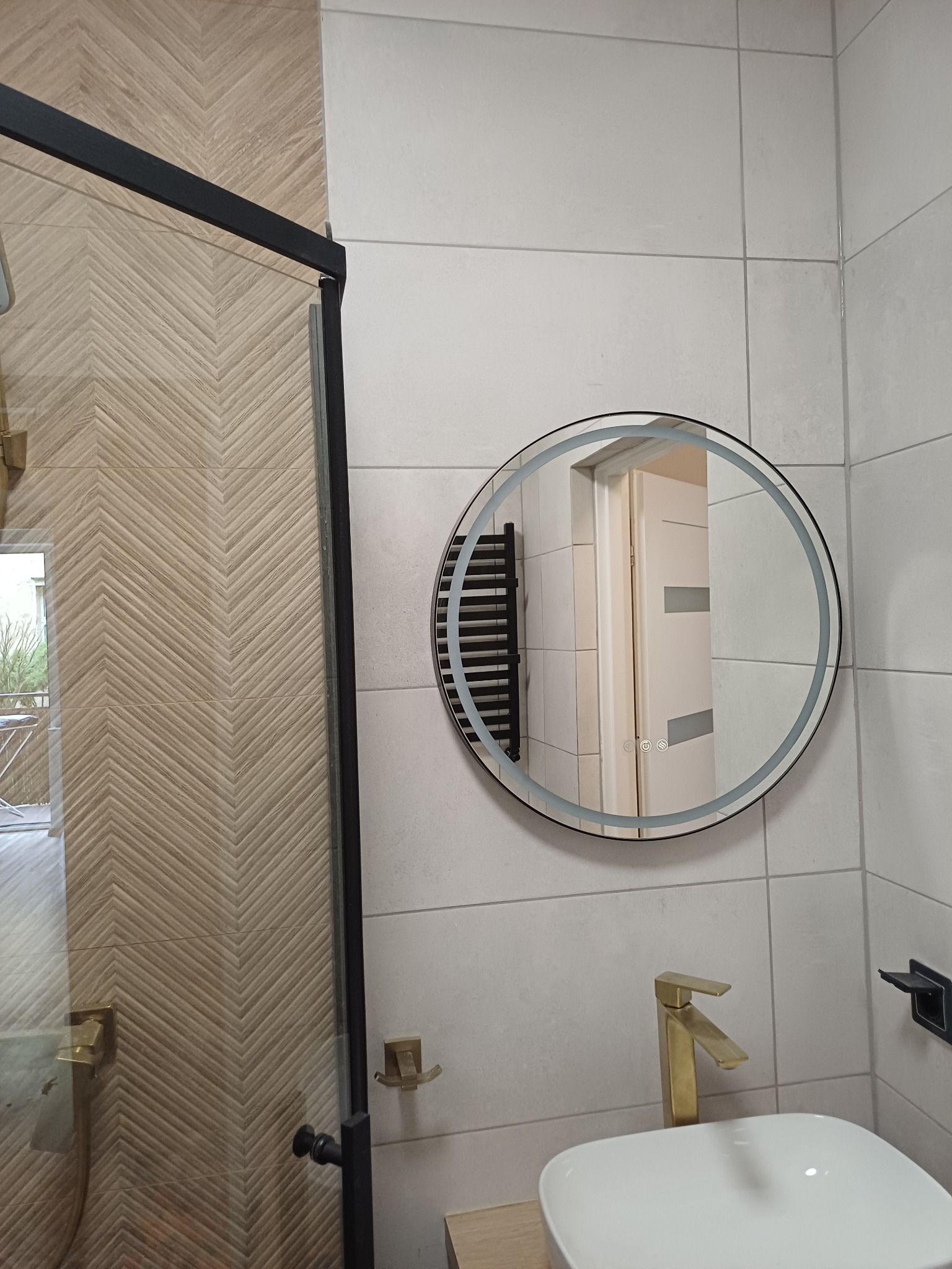 Nowoczesna łazienka z kabiną prysznicową, okrągłym lustrem z podświetleniem LED i złotą armaturą. Płytki w odcieniach bieli i beżu tworzą spójną całość.