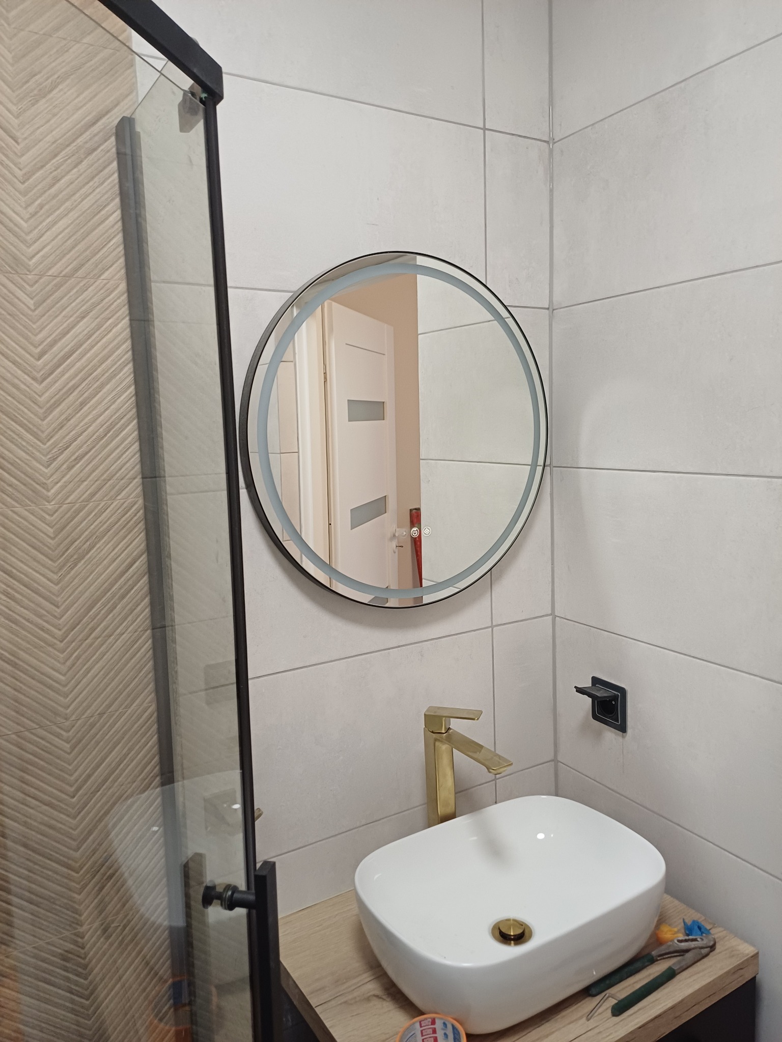 Nowoczesna łazienka w Człopie: okrągłe lustro z podświetleniem, biała umywalka nablatowa z złotą baterią, płytki w odcieniach szarości i kabina prysznicowa.