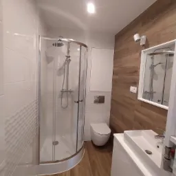 Nowoczesna łazienka z kabiną prysznicową typu walk-in, umywalką nablatową na białej szafce, wiszącą toaletą i lustrem z oświetleniem, wykończona płytkami imitującymi drewno na jednej ścianie...