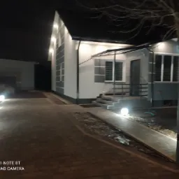 Nocne ujęcie nowoczesnego domu z oświetloną elewacją i ścieżką z kostki brukowej, oświetloną kulistymi lampami.