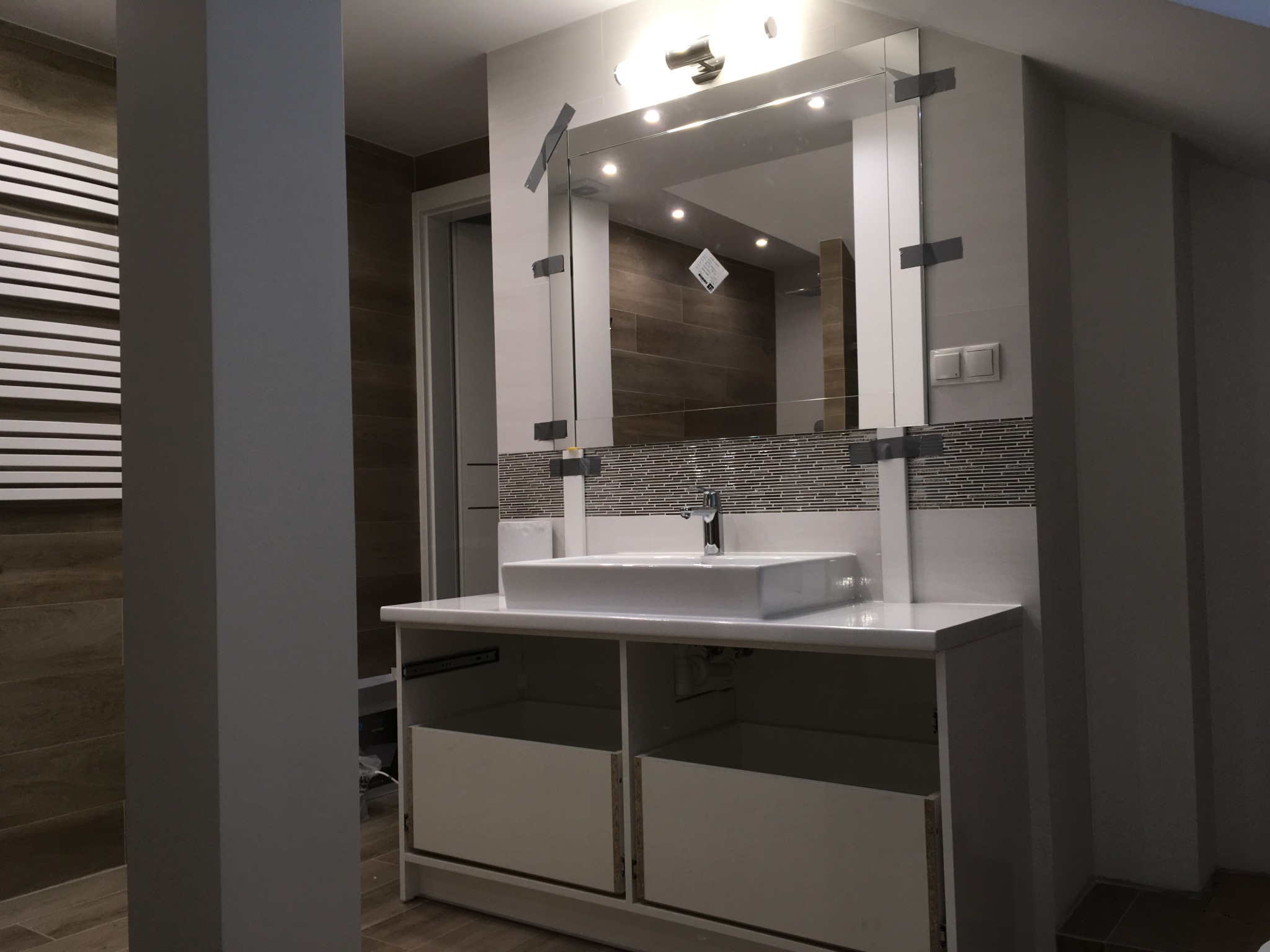 Nowoczesna łazienka w trakcie wykańczania: biała prostokątna umywalka nablatowa na szafce z wysuwanymi szufladami, duże lustro z taśmą montażową, płytki mozaikowe nad umywalką.