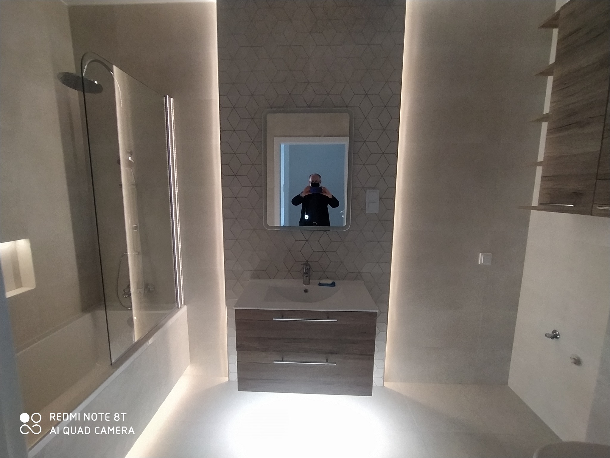 Nowoczesna łazienka z podświetleniem LED, kabiną prysznicową, umywalką z szafką w odcieniach drewna i lustrem, w którym odbija się osoba robiąca zdjęcie.