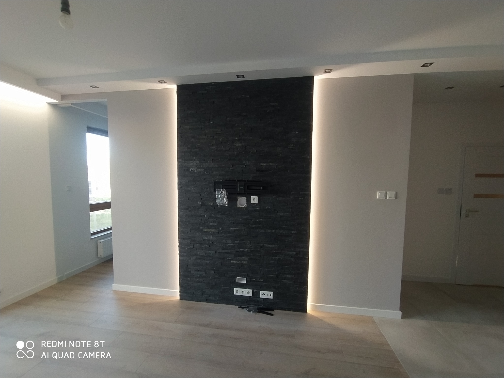 Salon po metamorfozie z czarną ścianą z kamienia dekoracyjnego, podświetloną po bokach, z przygotowanymi gniazdkami i uchwytem pod telewizor, z widocznym fragmentem podłogi i sufitu z oświetleniem...