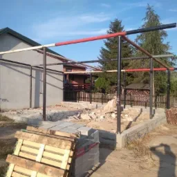 Konstrukcja metalowa z rur, częściowo pomalowana na czerwono i czarno, w trakcie budowy z fundamentami z betonowych bloczków, widoczne palety i gruz.