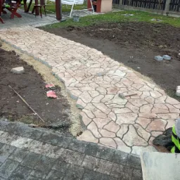 Ścieżka z nieregularnych płyt kamiennych w trakcie układania, widoczny młotek, rozsypany żwir, fragment ogrodzenia i altany w tle.