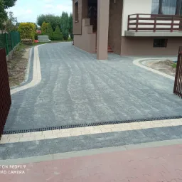 Szeroki podjazd z szarej kostki brukowej z jasnym obramowaniem, zakończony liniowym odpływem, prowadzący do domu z beżową elewacją i drewnianą balustradą, widoczne fragmenty ogrodzenia.