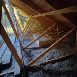 Poddasze z drewnianą konstrukcją więźby dachowej wypełnione szarą izolacją w postaci wełny mineralnej, widoczne fragmenty desek poszycia dachu.